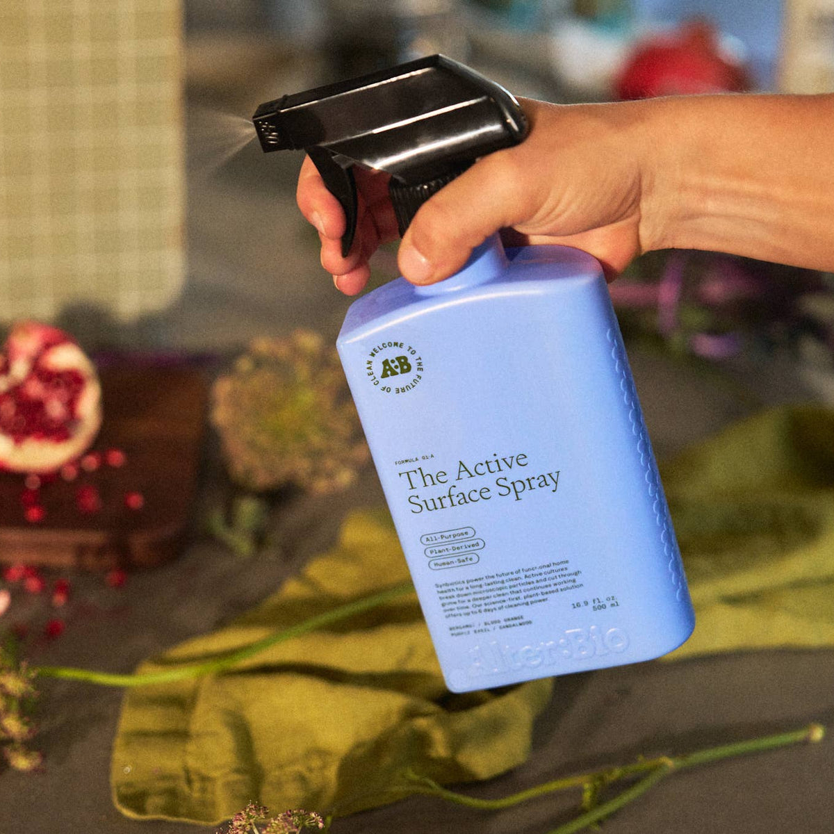 Hand holding a blue spray bottle labeled 'The Active Surface Spray' with a blurred background of vegetables and flowers.