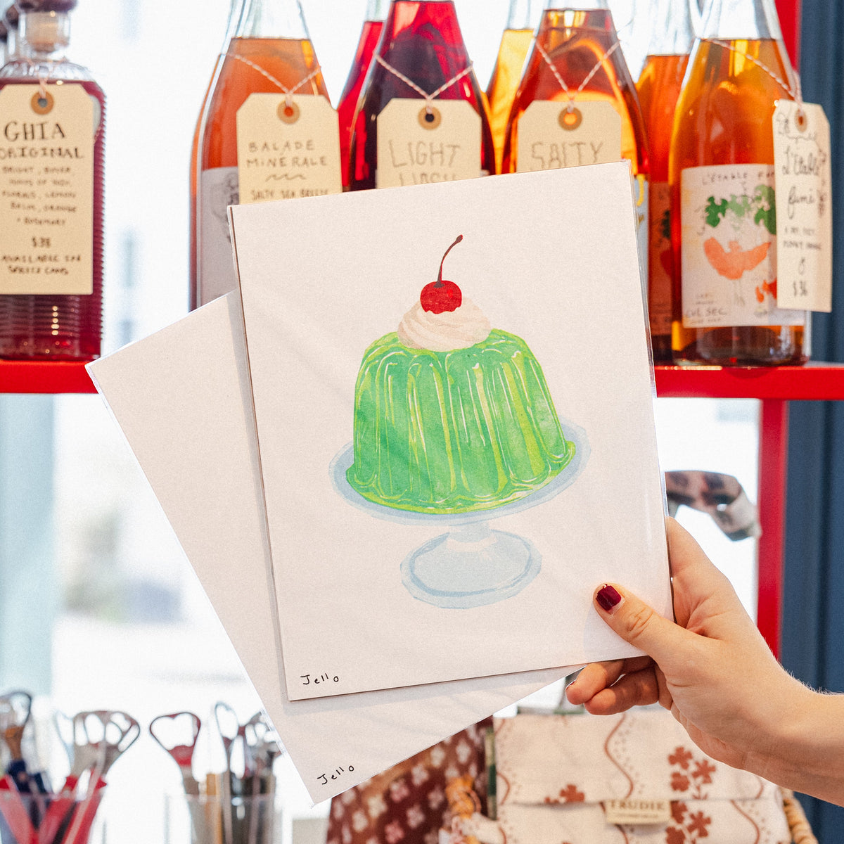 A hand holds a white art print featuring a hand-drawn green jelly bean dessert with a red cherry, set against a red shelving unit filled with various glass bottles.