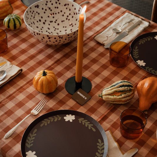 A styled dining table setting featuring dark brown ceramic plates with a white floral wreath border, a speckled cream bowl, amber glass tumblers, and a lit orange taper candle on a black holder. The table is covered with a rust-orange and beige gingham checkcloth and decorated with small pumpkins and gourds.