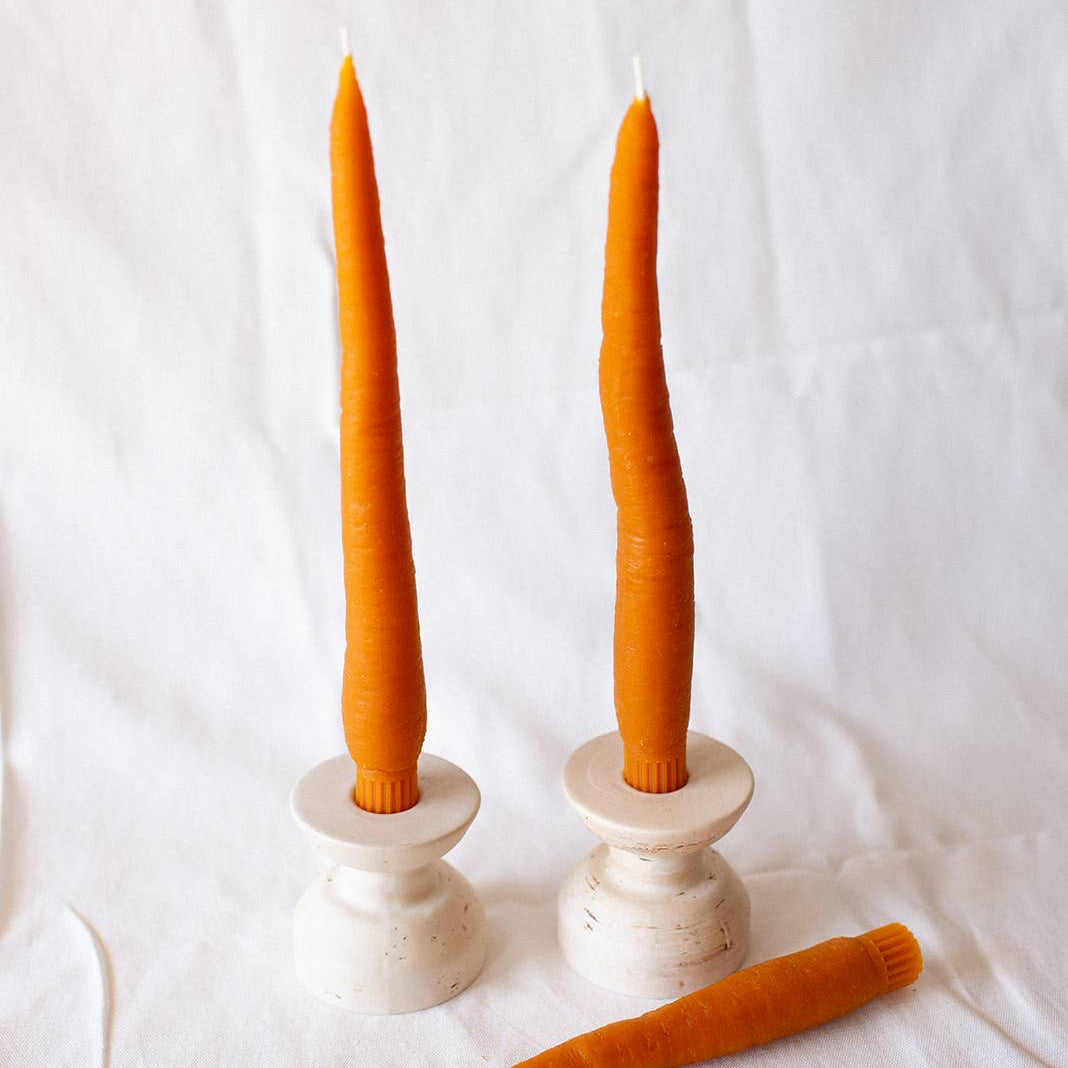 Two carrot-shaped taper candles on white candle holders, with one upright and one lying on its side, against a textured white background.