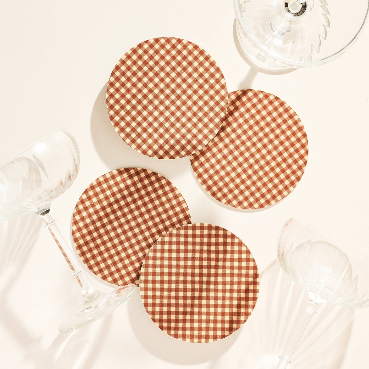 Four round coasters with a brown and beige gingham checkered pattern, arranged on a light cream background with wine glasses around them.