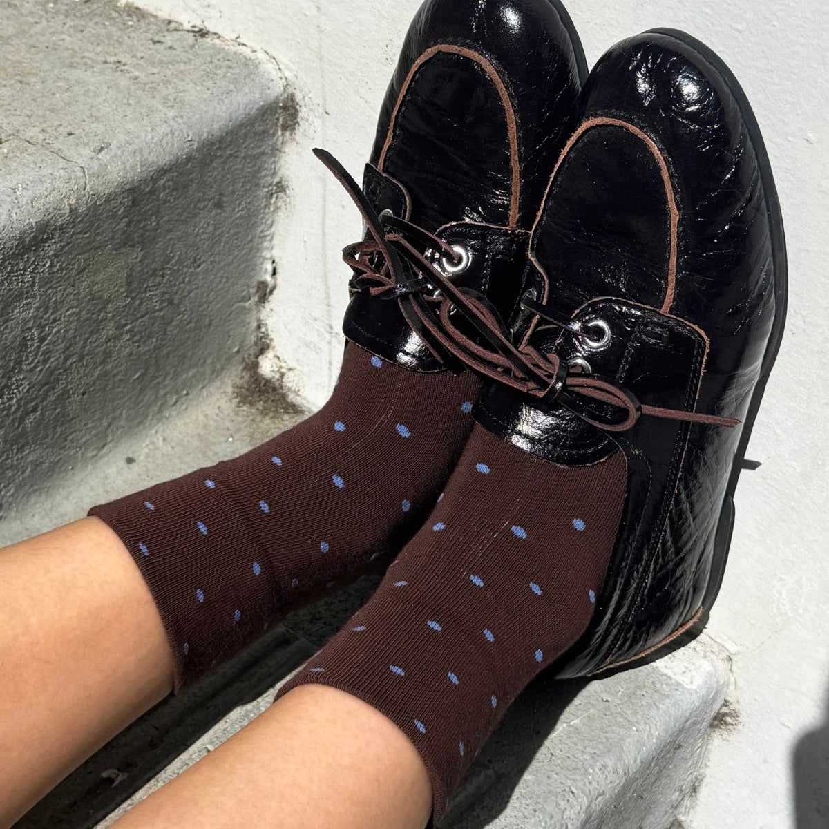 A person sitting on concrete steps wearing dark brown boat shoes with brown laces and matching dark brown socks with small blue polka dots.