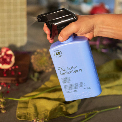 Hand holding a blue spray bottle labeled 'The Active Surface Spray' with a blurred background of vegetables and flowers.