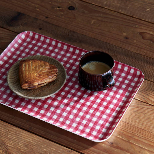 A red and white checkered linen tray placed on a wooden surface, with a cup of coffee and a plate with a pastry on it.