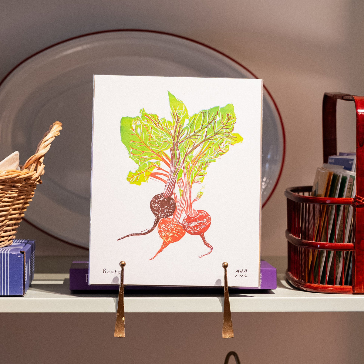 A styled bookshelf display featuring a white art print with a colorful illustration of a beet and greens, displayed on a small easel. The surrounding shelves contain various home decor items, including a wicker basket, a red book basket, red 'Cookbooks' boxes, and other decorative objects.