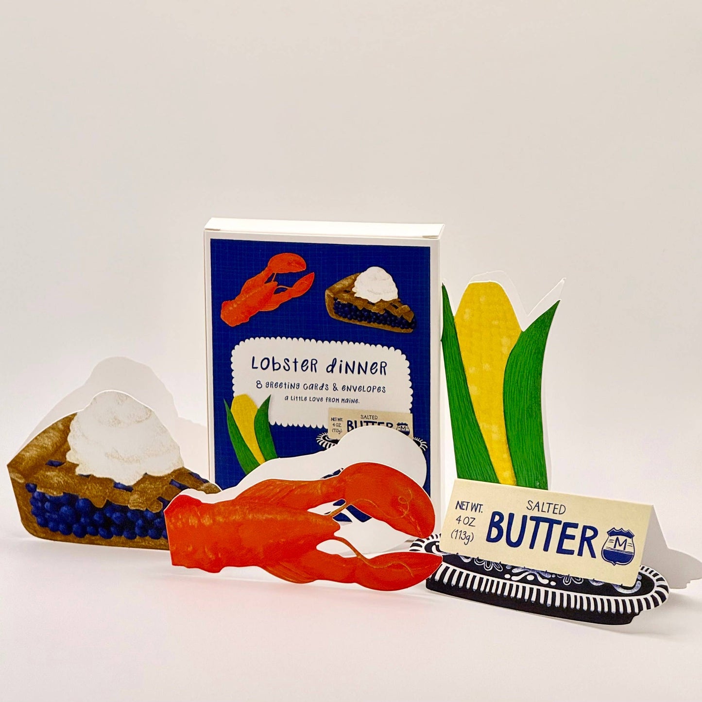 A set of paper craft cards and a small butter-style stick for a lobster dinner themed party, displayed on a white background. The set includes a blue 'Lobster Dinner' card, a red lobster claw, a yellow corn card, a pie card, and a novelty butter stick with blue and white packaging.