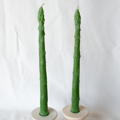 A pair of green asparagus-shaped beeswax candles on white candle holders against a white background.