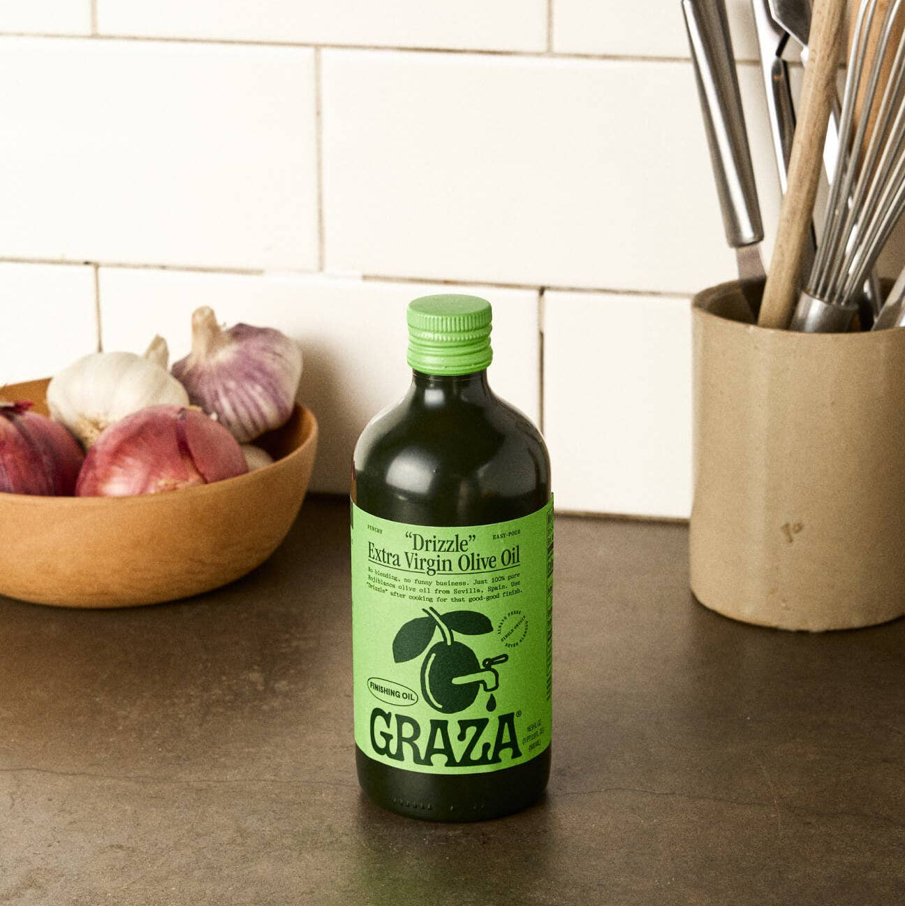Bottle of Graza olive oil on a kitchen counter with ingredients and utensils in the background.
