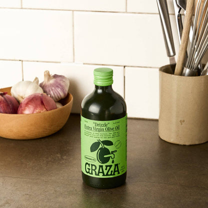Bottle of Graza olive oil on a kitchen counter with ingredients and utensils in the background.