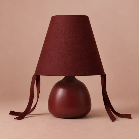 A table lamp with a tapered fabric shade in deep burgundy, featuring two long hanging ties, and a rounded glossy base in a matching dark red-brown finish, set against a solid warm beige background.